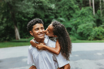 A little sister on her big teenage brother's back giving him a kiss on the cheek in an endeering moment amongst siblings in a family