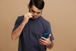 © SHOTPRIME STUDIO - handsome man with a phone in hand in a gray T-shirt and glasses tin hands beige background