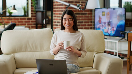 Poster - Asian woman sitting in living room working from home while smiling at camera. Happy student inside modern apartment having laptop on table while waiting for online class to start.