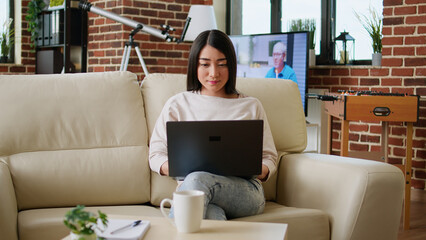 Poster - Focused young adult woman working remotely on laptop while sitting on sofa in living room at home. Attractive asian freelancer doing remote work on modern portable computer.