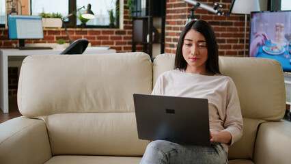 Poster - Focused woman sitting on sofa while working remotely on laptop. Beautiful young adult asian student attending online class while typing important lessons on modern portable computer at home.