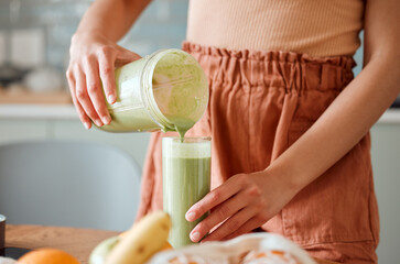 Wall Mural - Woman pouring healthy smoothie in a glass from a blender jar on a counter for detox. Female making fresh green fruit juice in her kitchen with vegetables and consumables for a fit lifestyle.