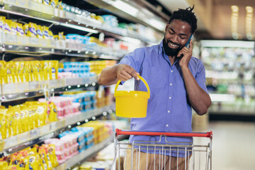 Black Male Buyer Shopping Groceries In Supermarket Taking Product From Shelf Standing With Shop Cart Indoors.