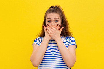 Wall Mural - Scared girl in white-blue striped t shirt covers mouth hands and looking at camera with shocked facial expression. Young emotional woman