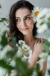 Smiling woman with visage and chamomile in hair looking away near blurred plants isolated on grey.
