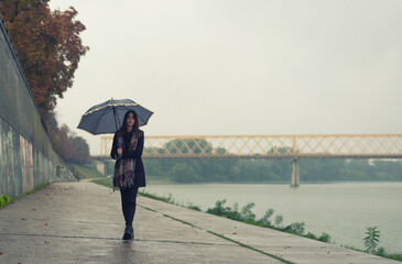 Wall Mural - Young woman with umbrella walking in the autumn rain beside river