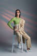 Stylish nonbinary model posing while sitting on chair on abstract background.