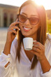 © Paulista - Smiling young woman with sunglasses holding mobile phone and enjoying a cup of coffe outdoor.