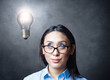 © Paulista - Pensive young woman with eyeglasses looking up to lightbulb light on concrete wall background. Creative thinking concept.