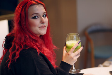 Wall Mural - European plus size woman drink green smoothie in cafe. Young red pink haired body positive girl sitting at restaurant