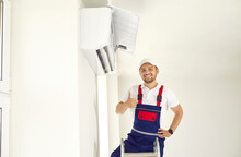 Happy Young Man In Uniform Standing On Ladder Under New Air Conditioner Unit In White Room, Smiling And Showing Thumbs Up After A Job Well Done. AC Installation, Maintenance And Repair Service Concept