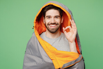 Young happy fun traveler white man wrapped in sleeping bag show ok isolated on plain green background. Tourist leads active healthy lifestyle walk on spare time. Hiking trek rest travel trip concept.