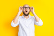 Bald young man holding glasses looks up in surprise on a yellow background