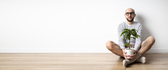 Young man in glasses holding potted flower while sitting on wooden floor. Banner.