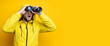 Surprised young man in yellow raincoat looking through binoculars on yellow background. Banner
