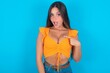 beautiful brunette woman wearing orange tank top over blue background being in stupor shocked, has astonished expression pointing at oneself with finger saying: Who me?