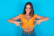 © Jihan - beautiful brunette woman wearing orange tank top over blue background with positive expression, points down with both index fingers, has broad interested smile. Look there, please.