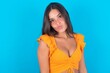 beautiful brunette woman wearing orange tank top over blue background , keeps lips as going to kiss someone, has glad expression, grimace face. Standing indoors. Beauty concept.
