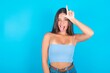 beautiful brunette woman wearing blue tank top over blue background gestures with finger on forehead makes loser gesture makes fun of people shows tongue