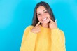 Happy beautiful brunette woman wearing yellow sweater over blue background with toothy smile, keeps index fingers near mouth, fingers pointing and forcing cheerful smile