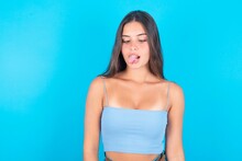 Funny Beautiful Brunette Woman Wearing Blue Tank Top Over Blue Background Makes Grimace And Crosses Eyes Plays Fool Has Fun Alone Sticks Out Tongue.