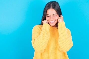 beautiful brunette woman wearing yellow sweater over blue background grins joyfully, imagines something pleasant, copy space. Pleasant emotions concept.