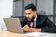 © Kateryna - Shocked puzzled stunned indian or arabian young businessman, company ceo, in a suit, sits in modern office, surprised look at laptop screen, see unexpected message or news, surprised facial expression