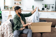 © Kateryna - Frustrated arabian or indian man in casual clothes, sitting on the sofa in the living room, unpacking his parcel, taking out clothes from the cardboard box, looking at her disappointedly, dissatisfied