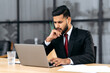 © Kateryna - Confused puzzled indian or arabian young businessman, entrepreneur, in a suit, sitting in his modern office, focused look at laptop screen, see unexpected message or news, is under stress