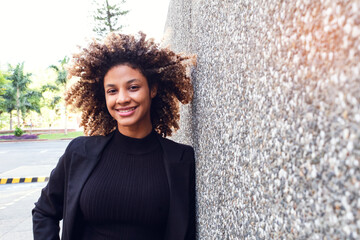 portrait of beautiful elegant african american adult woman smiling at camera leaning on wall in city outdoors happy on sunny day.