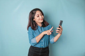 Wall Mural - A portrait of a happy Asian woman is smiling and holding her smartphone wearing a blue shirt isolated by a blue background