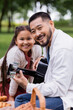 © LIGHTFIELD STUDIOS - Positive asian man playing acoustic guitar and looking at camera near daughter in park.