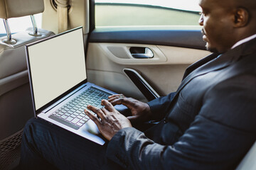 Wall Mural - Business concept of a handsome dark-skinned man in a suit and glasses in the back seat of a luxury car working with a laptop. Copyspace and mockup