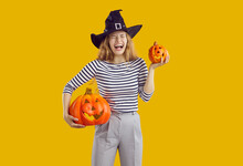 Young Girl Having Fun On Halloween Night. Studio Shot Of A Happy, Joyful, Funny Woman Wearing A Black Witch Hat Standing Isolated On A Yellow Background, Holding Two Orange Pumpkins And Laughing