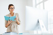 © VK Studio - Young dark haired woman in elegant wear holds blue notepad focused into monitor, ponders about business strategy for eneterprise, wears transparent eyeglasses, poses in spacious white own cabinet