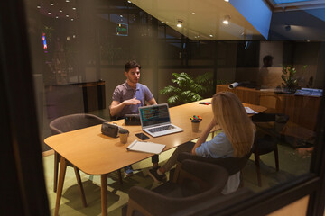 Through glass shot of male and female software engineers or programmers discussing project in meeting in office, laptop with code on screen and vr headsets on desk