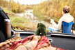 © Syda Productions - travel, tourism and camping concept - view to river from car trunk with feet in warm socks under blanket, two cups of tea, thermos and cookies