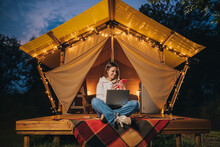 Happy Woman Freelancer Using Phone And Working Laptop On Cozy Glamping Tent In Summer Night. Luxury Camping Tent For Outdoor Holiday And Vacation. Lifestyle Concept