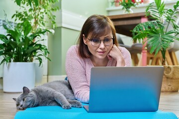 Wall Mural - Middle-aged woman lying with cat on exercise mat, looking at laptop