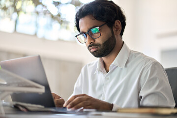 Busy indian business man office employee worker, arab professional company manager analyst wearing eyeglasses working on corporate data typing on laptop computer sitting at home office work desk.