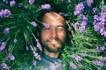 Portrait of young handsome bearded man lying among lavender flowers in blossom field. Happy smiling guy relax on the grass on sunny summer day. Freedom, love nature concept.