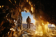  A Man Stands With His Back To The Cave And Admires Nature. View From The Ice Cave With Huge Icicles On The Frozen Lake Baikal At Sunset. Winter Travel Or Freedom Concept. 