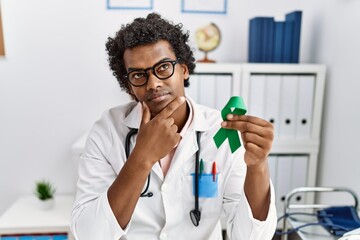 Poster - African doctor man holding support green ribbon serious face thinking about question with hand on chin, thoughtful about confusing idea