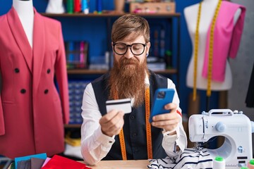 Wall Mural - Young redhead man tailor using smartphone and credit card at clothing factory