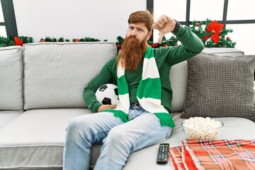 Canvas Print - Redhead man with long beard football hooligan holding ball sitting on the sofa with angry face, negative sign showing dislike with thumbs down, rejection concept