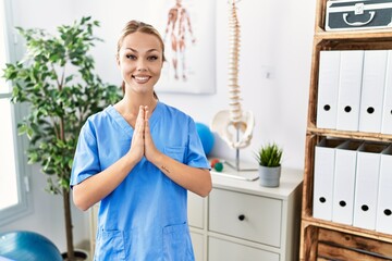 Sticker - Young caucasian woman working at pain recovery clinic praying with hands together asking for forgiveness smiling confident.