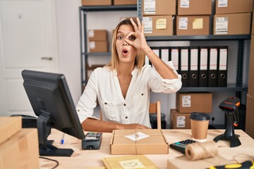 Poster - Young blonde woman working at small business ecommerce doing ok gesture shocked with surprised face, eye looking through fingers. unbelieving expression.