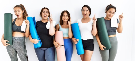 Group of women holding yoga mat standing over isolated background pointing finger up with successful idea. exited and happy. number one.