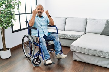 Wall Mural - Handsome senior man sitting on wheelchair at the living room celebrating surprised and amazed for success with arms raised and open eyes. winner concept.