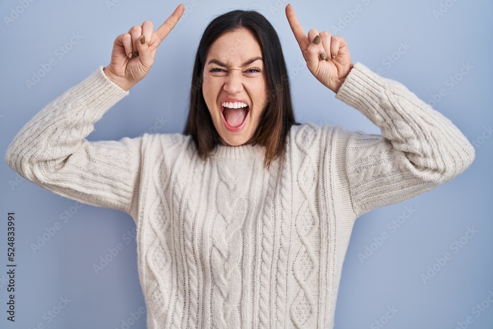 Young brunette woman standing over blue background smiling amazed and surprised and pointing up with fingers and raised arms.